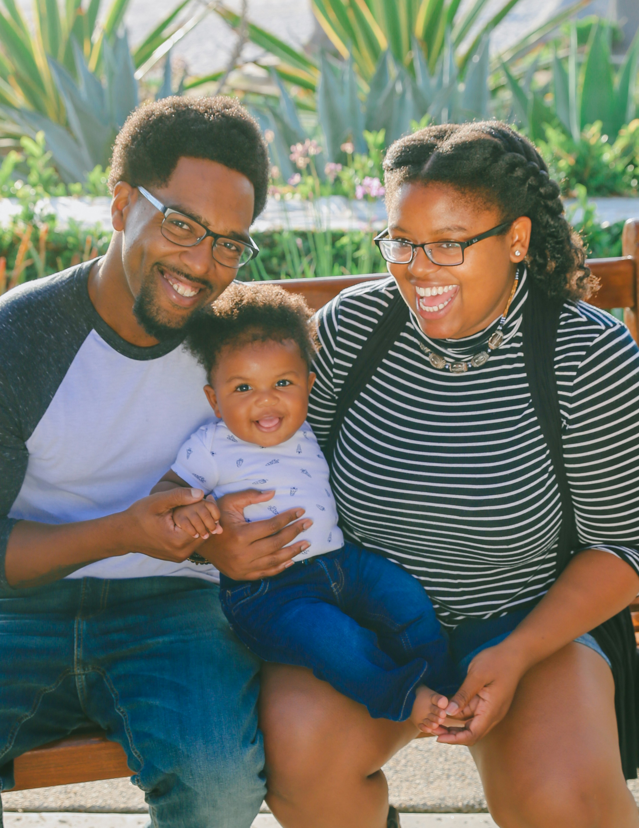 A young family — parents and their baby — sitting together on a bench outdoors, smiling