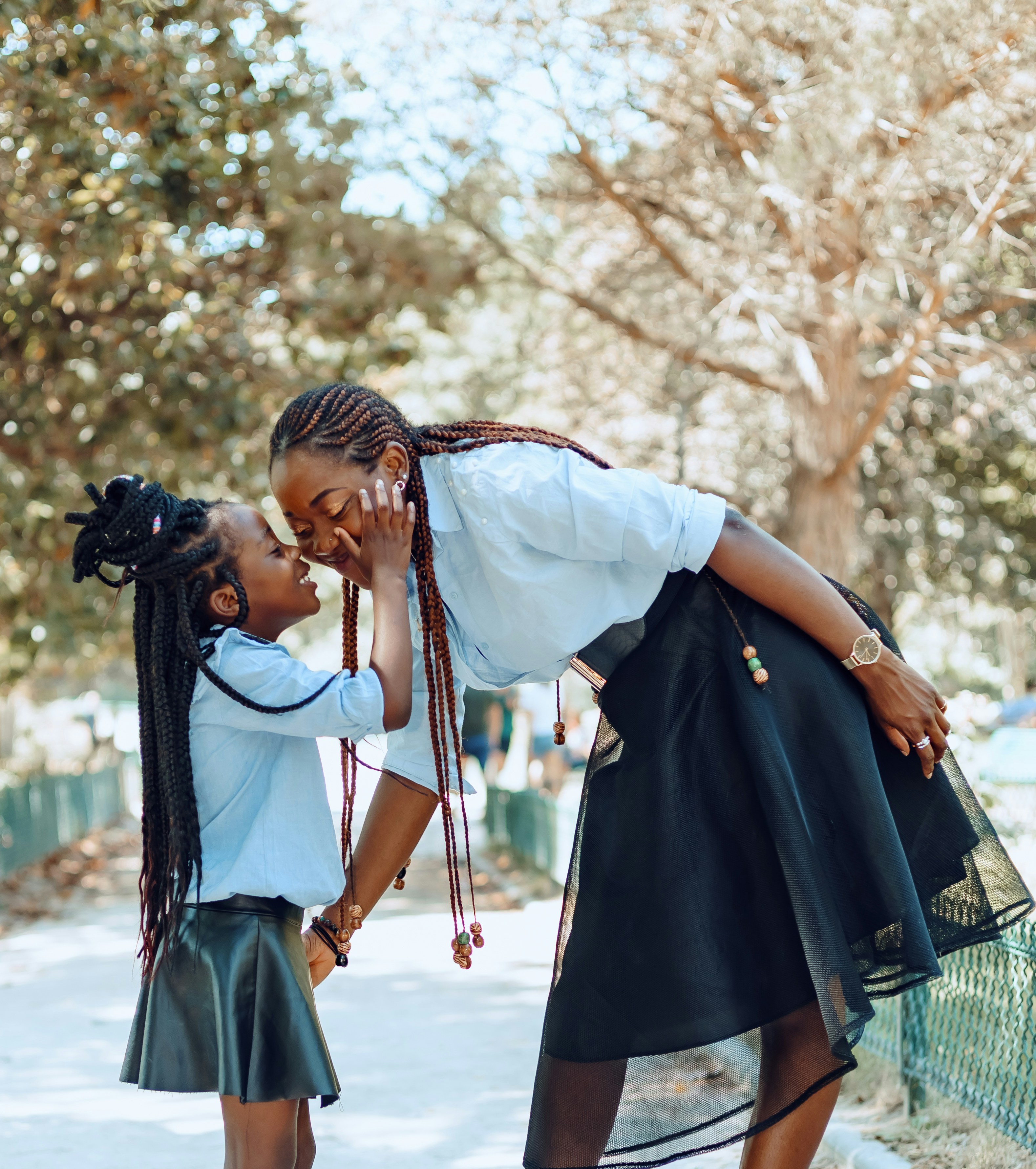 A mother leaning down to share a tender moment with her young daughter outdoors under trees